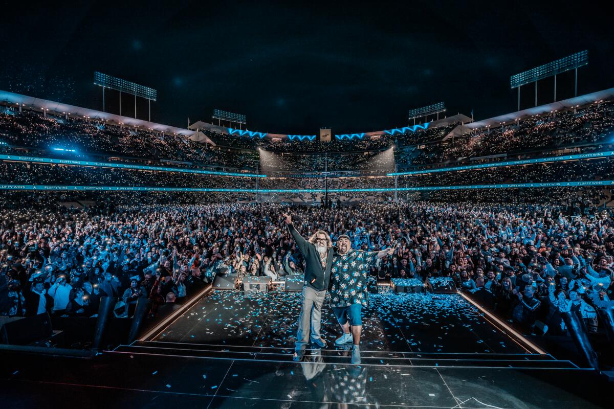 Two men stand in front of a sold out stadium crowd.