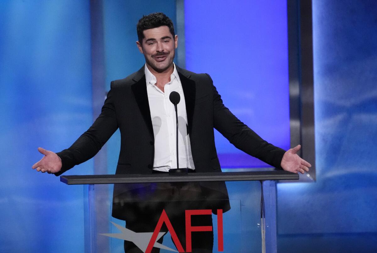 Zac Efron standing in a white shirt and dark blazer, his hands outstretched, behind a podium with the letters AFI on it