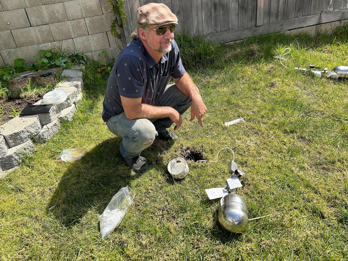 A man with a silver cannister and other scientific equipment squatting next to a small hole in a lawn.