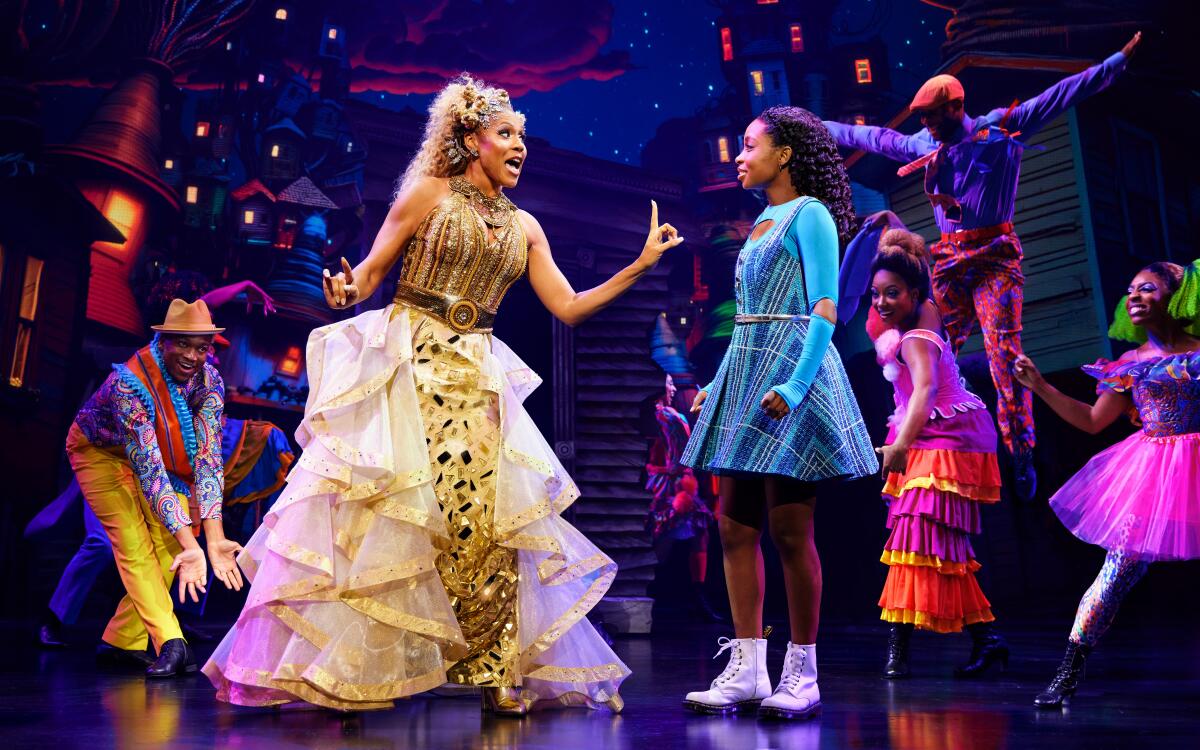 A woman in a gold dress makes a point to a girl in a blue dress on a colorful stage