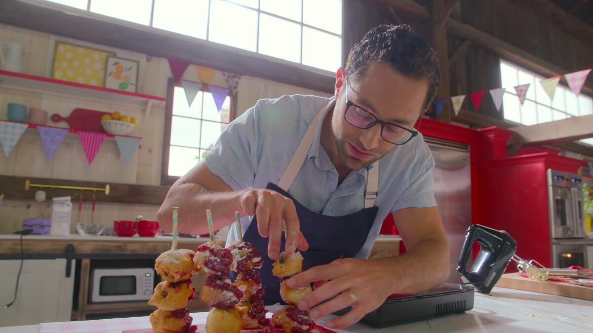 A man wearing a blue apron and glasses places a baked good on a stick.