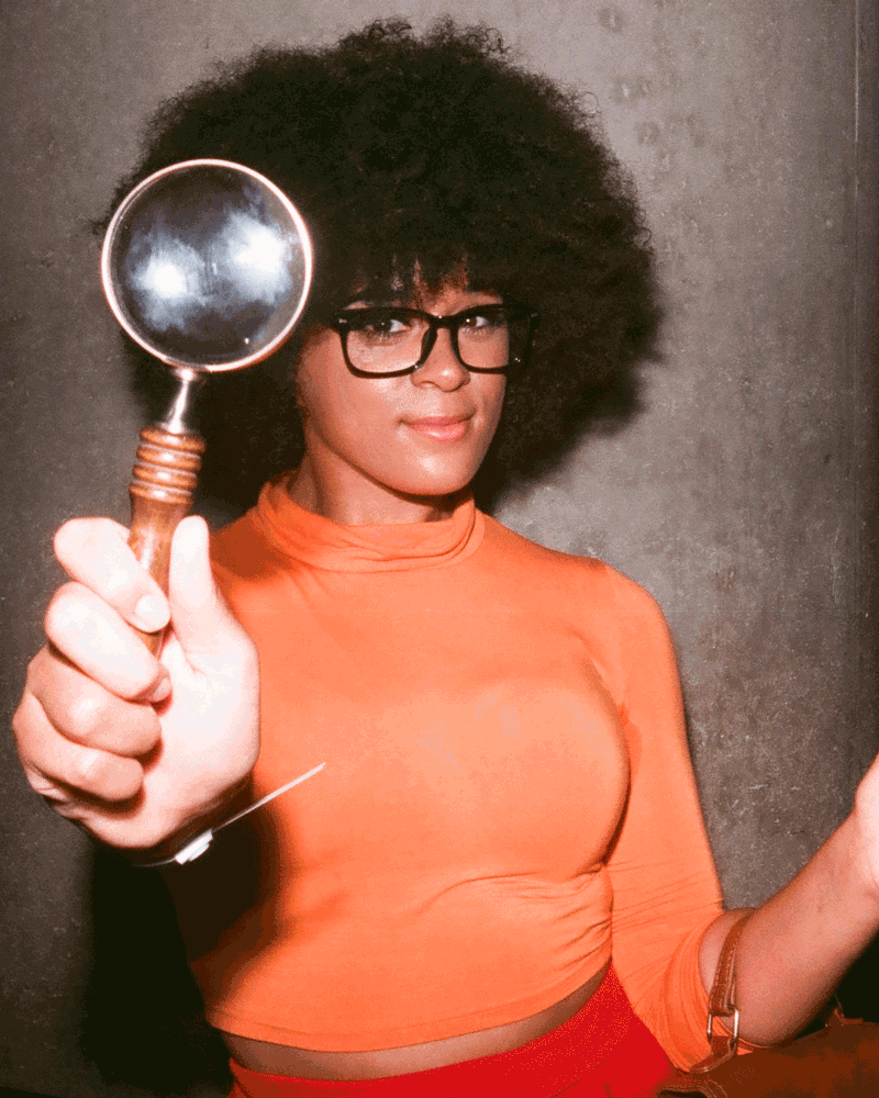 A gif of a woman in an orange turtleneck holding up a magnifying glass.