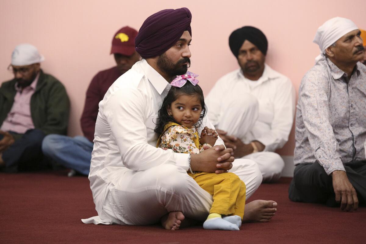 A man in a turban holds a little girl in his lap as he sits on a carpeted floor near several other men with head coverings