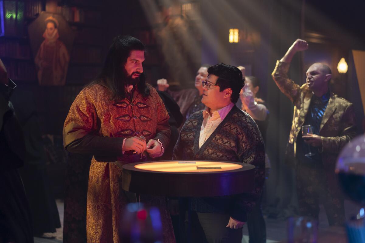 A bearded man dressed like an ancient talks with a younger man in a nightclub setting.