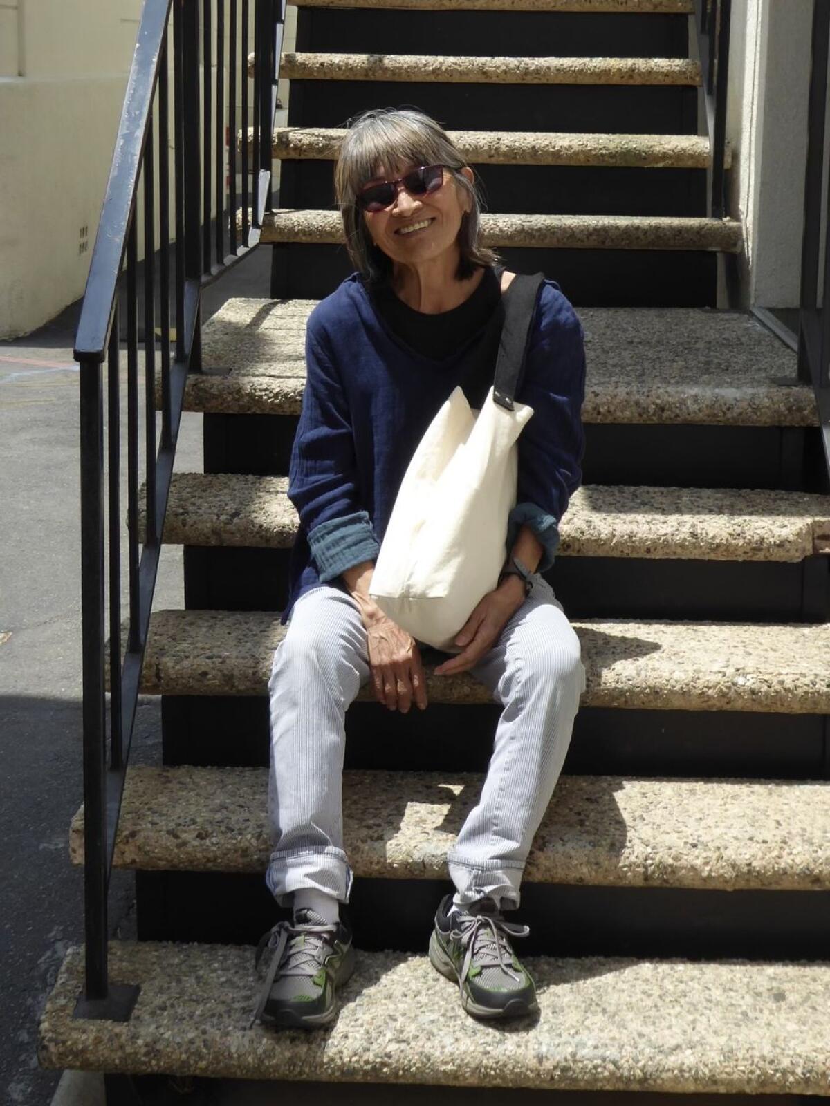 Eileen Saki wears sunglasses and smiling while sitting on steps and holding a tote bag