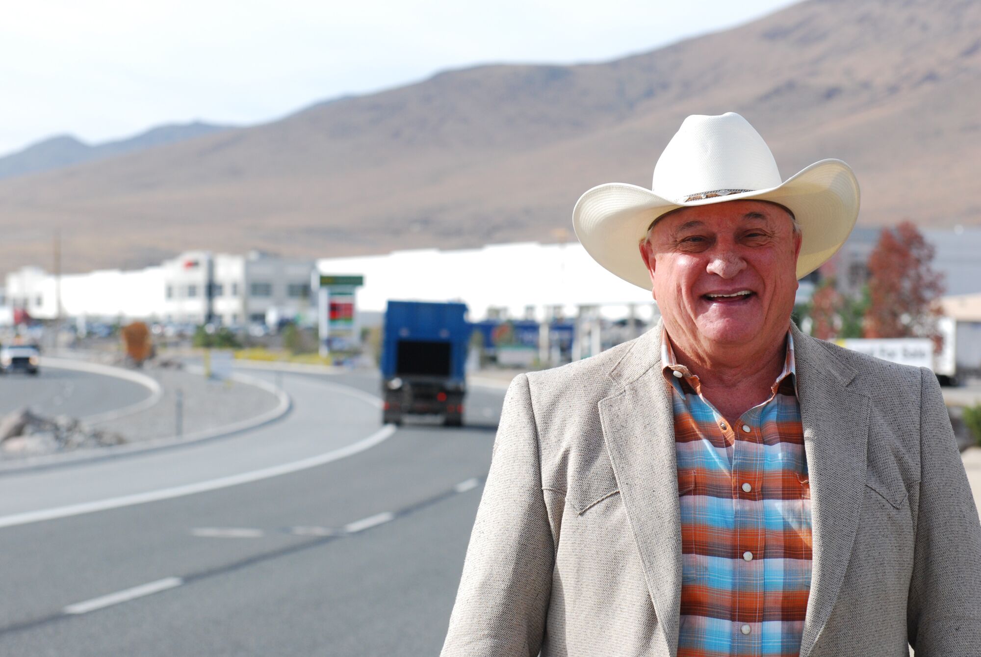 A man wearing a cowboy hat stands by a road and smiles.
