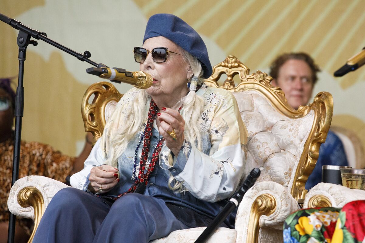 A woman with a beret and sunglasses sings while seated onstage