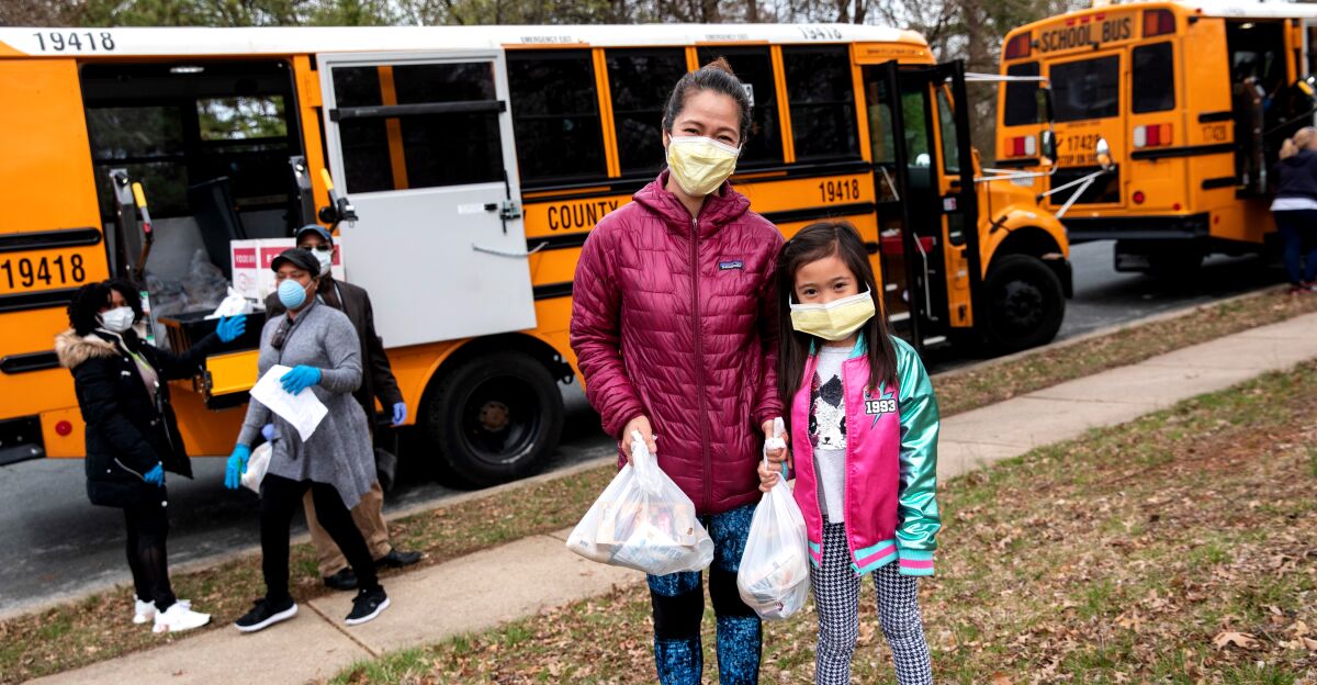 A woman and girl holding plastic bags stand in front of two yellow school buses