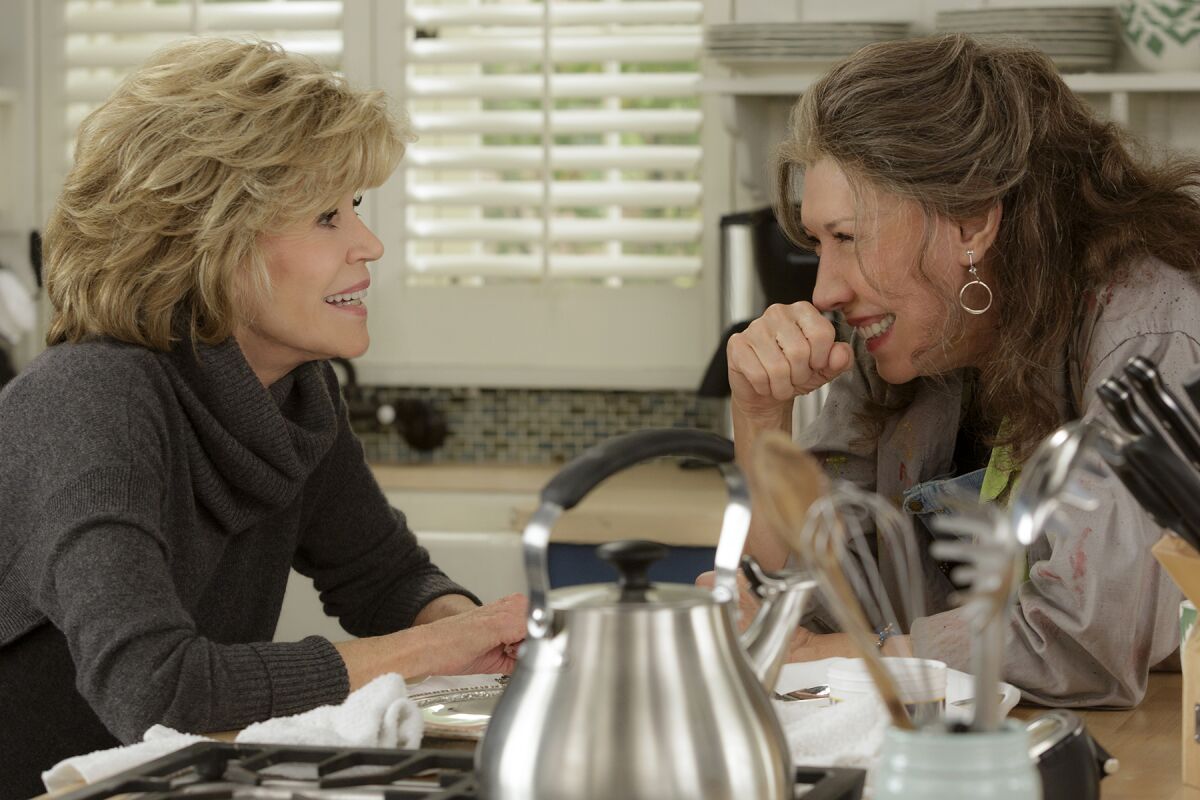 Jane Fonda and Lily Tomlin lean across a kitchen counter smiling at each other