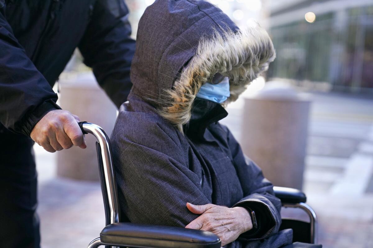 A woman is pushed in a wheelchair, her face obscured by the hood of her jacket.