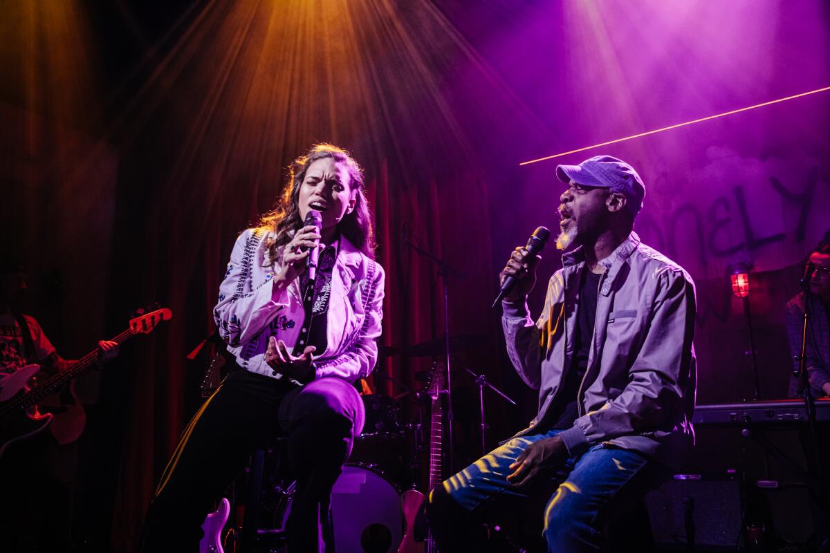 A man and a woman, seated onstage, sing into microphones