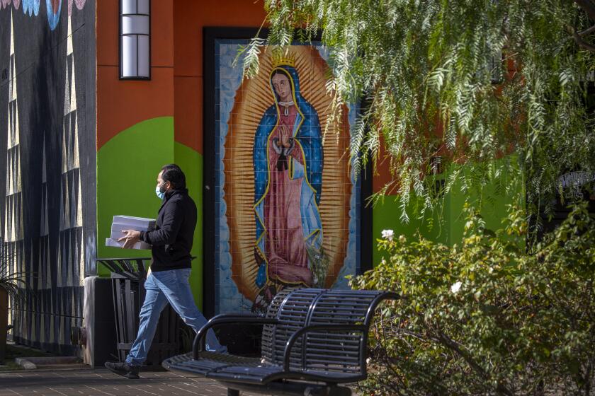 SANTA ANA, CA - NOVEMBER 18: A person wears a mask while carrying boxes of bakery good takeout and passing by a mural of Our Lady of Guadalupe at the Downtown Santa Ana Historic District Wednesday, Nov. 18, 2020. Orange County found itself back in the COVID-19 Purple Tier - the most restrictive in the state. (Allen J. Schaben / Los Angeles Times)