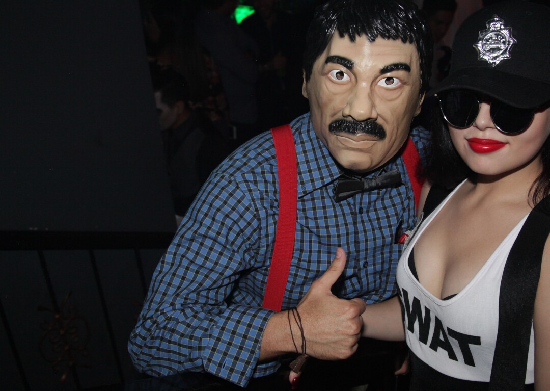 A snapshot-style images shows a masked man with a mustache next to a woman in sunglasses in a low-cut tank top.