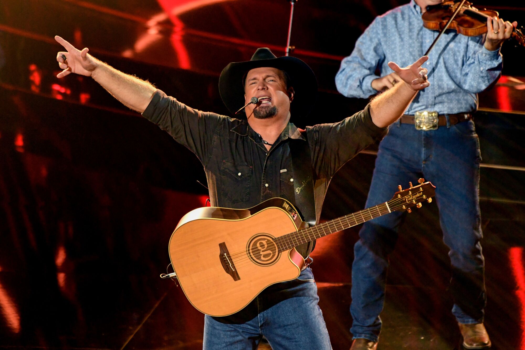 Garth Brooks, on stage with a fiddler, wears jeans and cowboy hat with his guitar slung over his shoulders. 