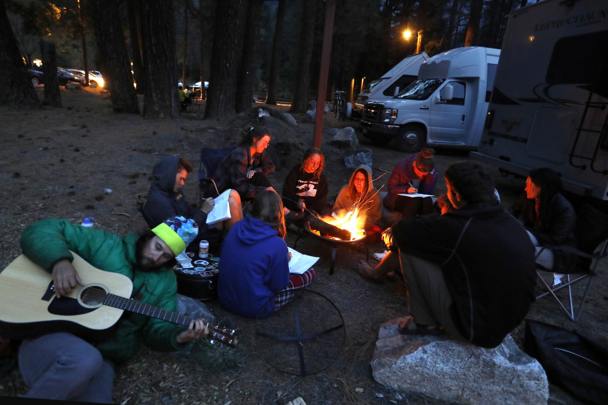 A man with a guitar lies against a rock as a group of teenagers surrounds a campfire
