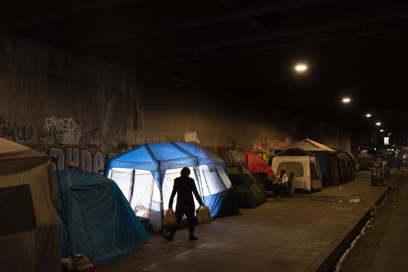 In this photo illuminated by an off-camera flash, a man walks past a homeless encampment underneath a bridge in Los Angeles, Thursday, Feb. 9, 2023. Homeless people in California are already a vulnerable group, often struggling with poor health, trauma and deep poverty before they lose their housing, according to a new study on adult homelessness. (AP Photo/Jae C. Hong)