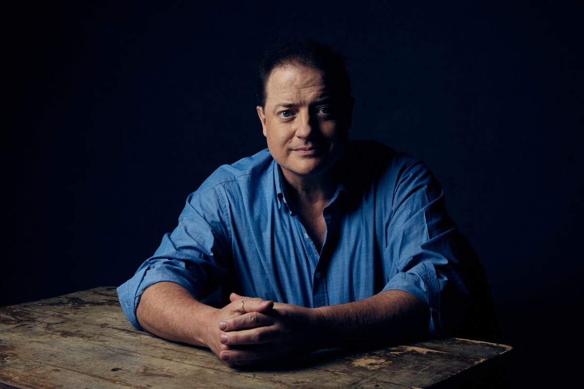 A portrait of Brendan Fraser in a blue shirt, leaning his arms on a table.