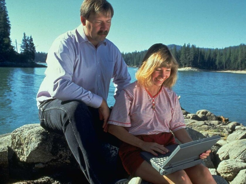 A man and a woman sit on some rocks looking at a vintage laptop computer, with a lake and trees behind them.