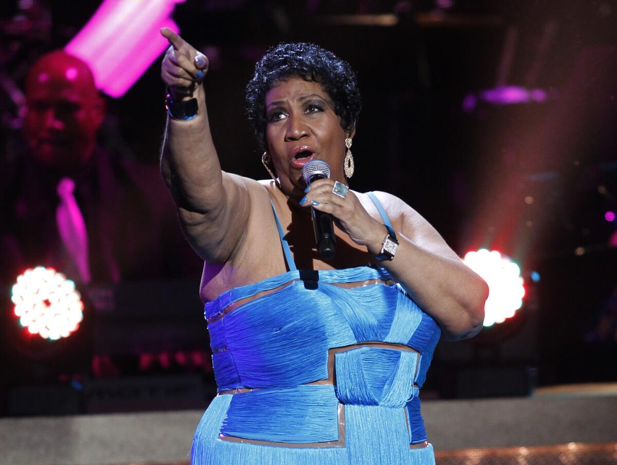 Aretha Franklin is pointing while holding a microphone to her mouth as she sings onstage, wearing a blue dress.