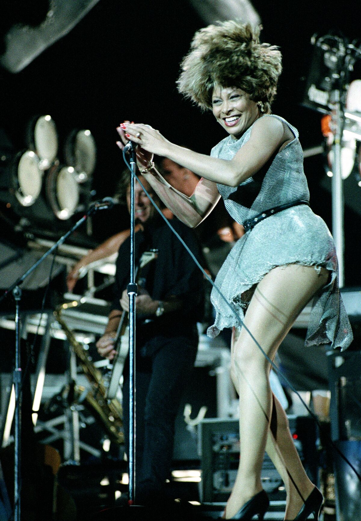 Tina Turner performing on stage in a short dress, smiling and holding a microphone stand with both hands. 