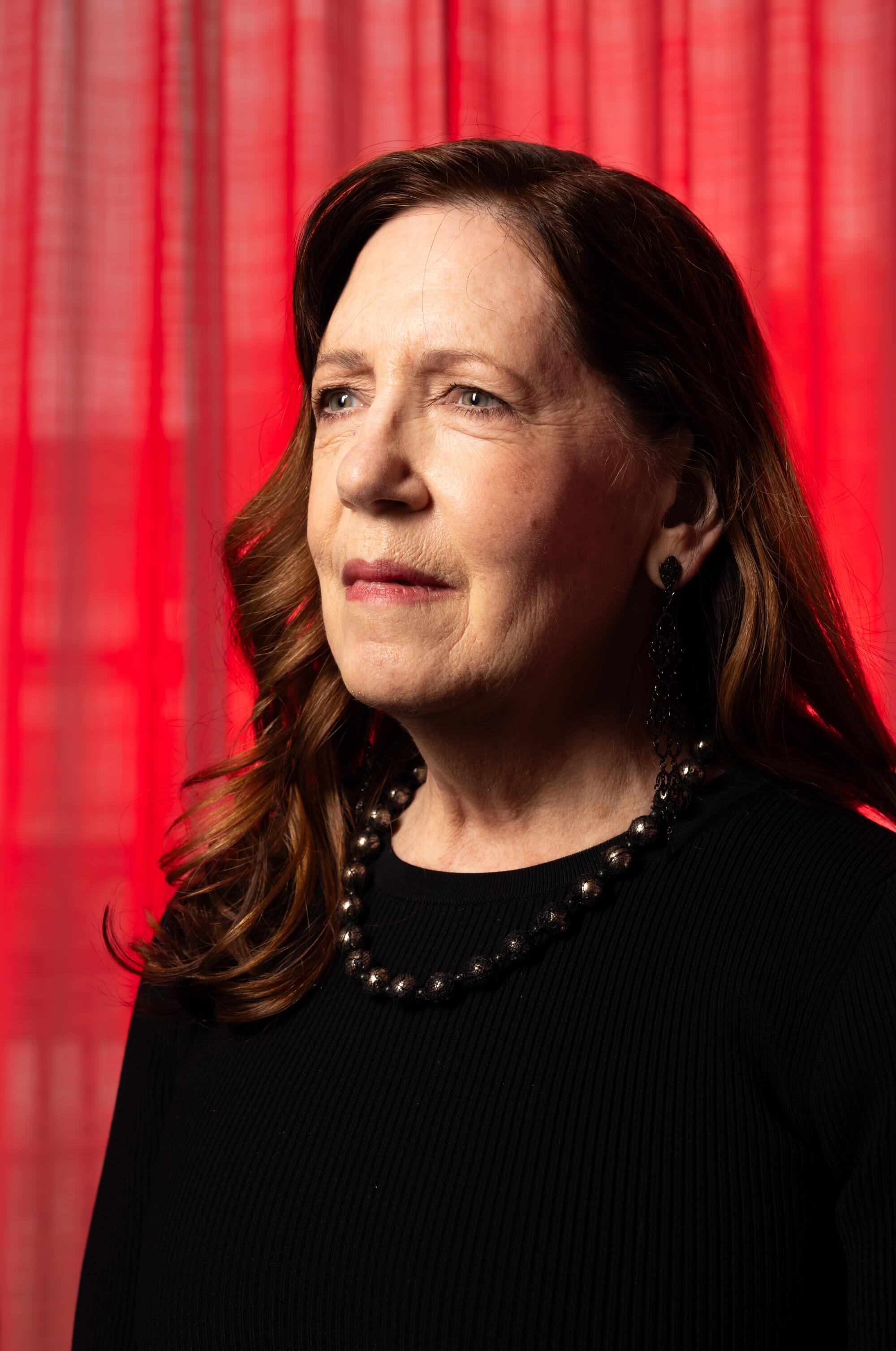 A woman with brown hair past her shoulders in a black top and necklace standing in front of a sheer red curtain.