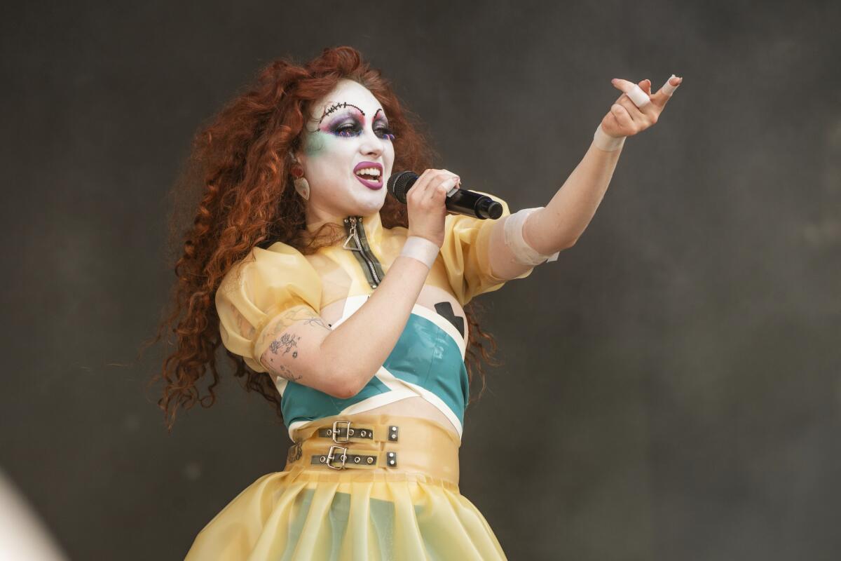 Chappell Roan raising one arm and singing into a mic onstage in long red curly hair and drag-style makeup