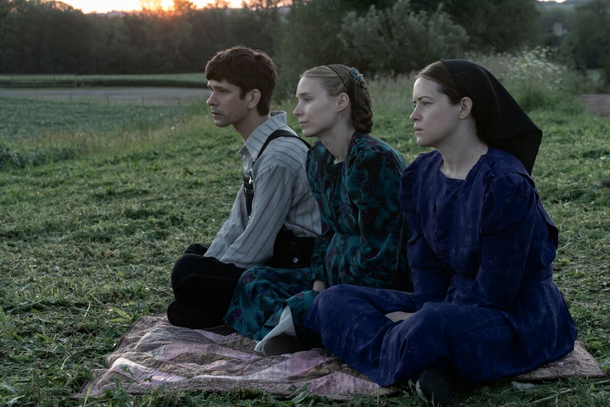 A man in overalls and two women in long dark dresses with their hair covered sit together on the grass