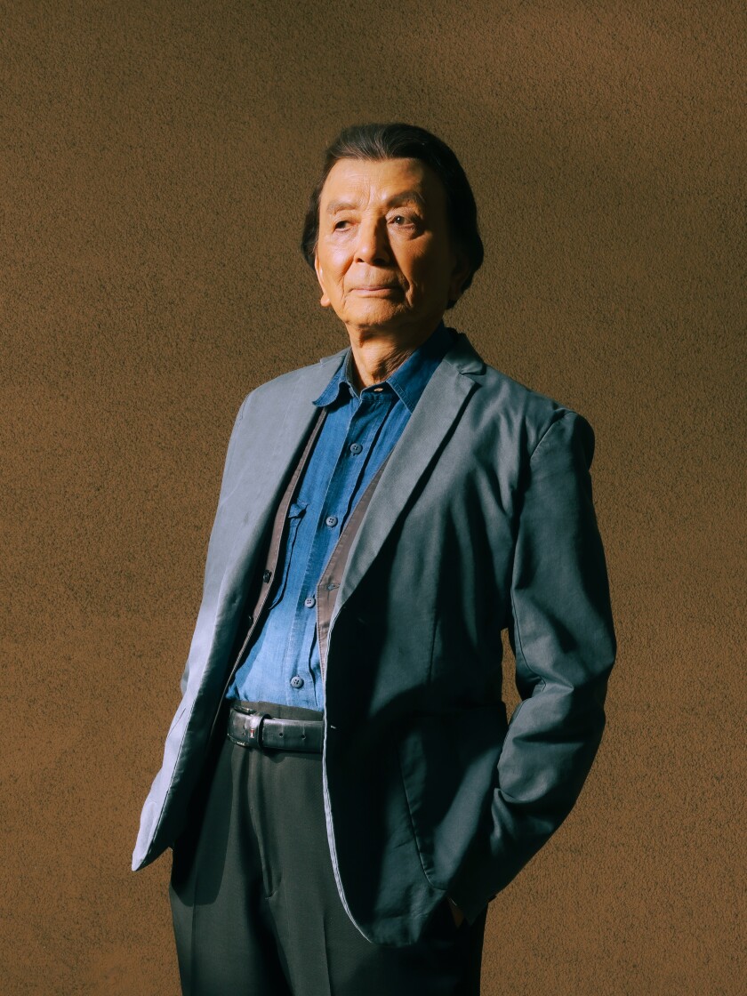 An older man in a suit stands in front of a brown background
