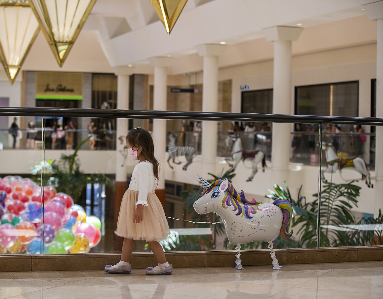 COSTA MESA, CA - JUNE 11: Arielle Wing, 3, of Newport Beach, pulls her balloon behind her while shopping with her mom, Aileen Wing, not pictured, as a long line of shoppers wait to enter Zara in the background as South Coast Plaza reopens, requiring customers maintain a social distance and wear face masks at South Coast Plaza Monday, June 11, 2020 in Costa Mesa, CA. Although not all stores in the upscale shopping center will resume operations, more than 110 merchants - including Apple, Macy's and Nordstrom - are opening their doors after being closed for three months amid the coronavirus pandemic. (Allen J. Schaben / Los Angeles Times)