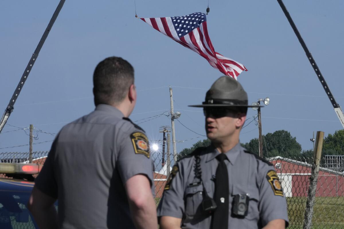 Police officers stand and talk, as a giant flag waves behind them, suspected between two cranes. 