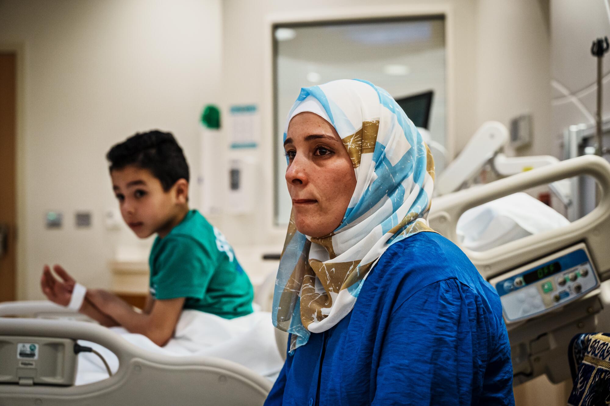 A woman in a white-and-blue patterned hijab and blue robe sits near a boy in a hospital bed 