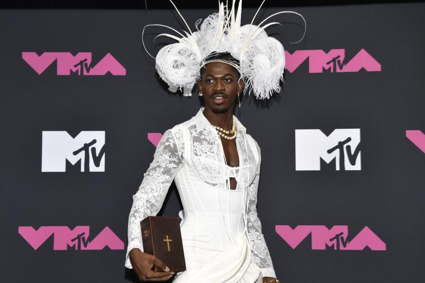 Lil Nas X poses in a white corset and feathered headdress holding a bible next to his right hip