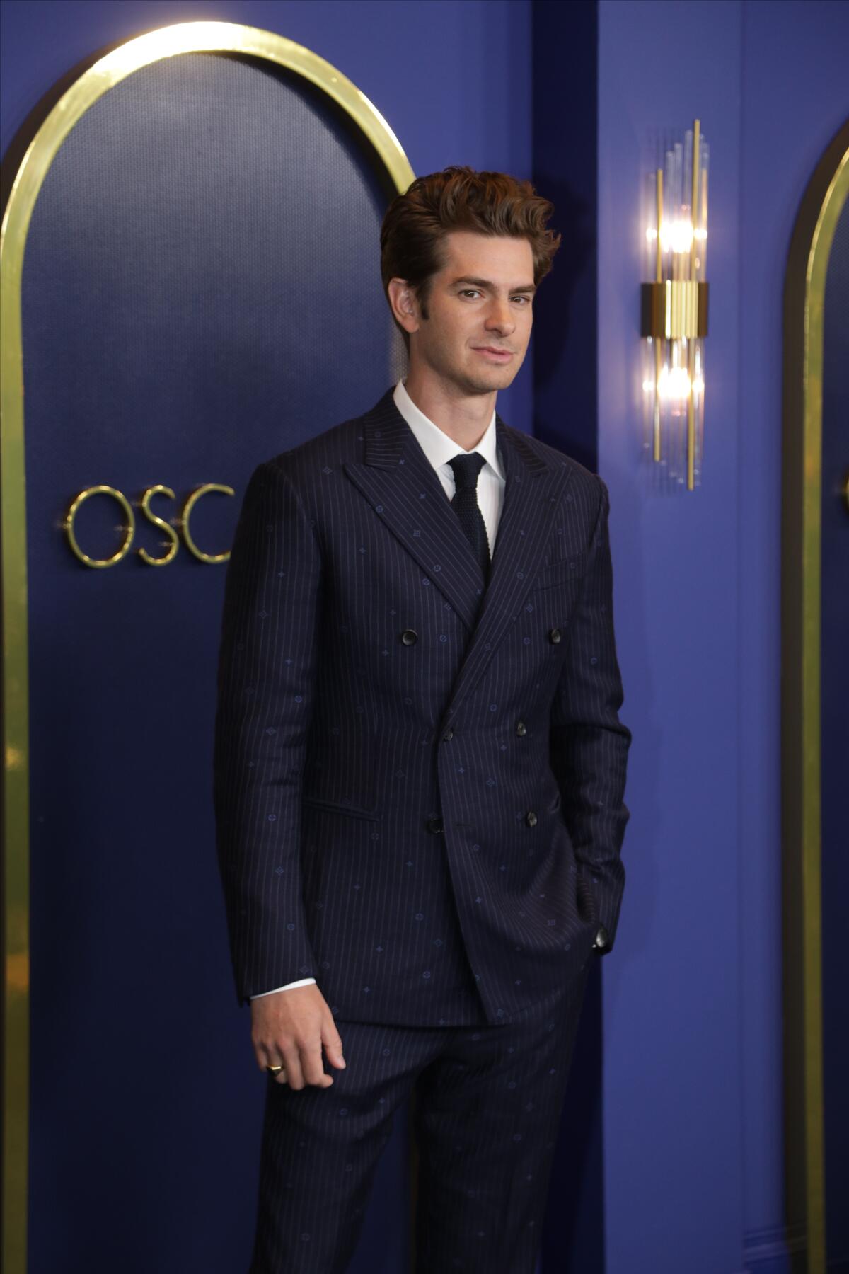 A man wearing a suit stands in front of a wall with the word Oscars on it.