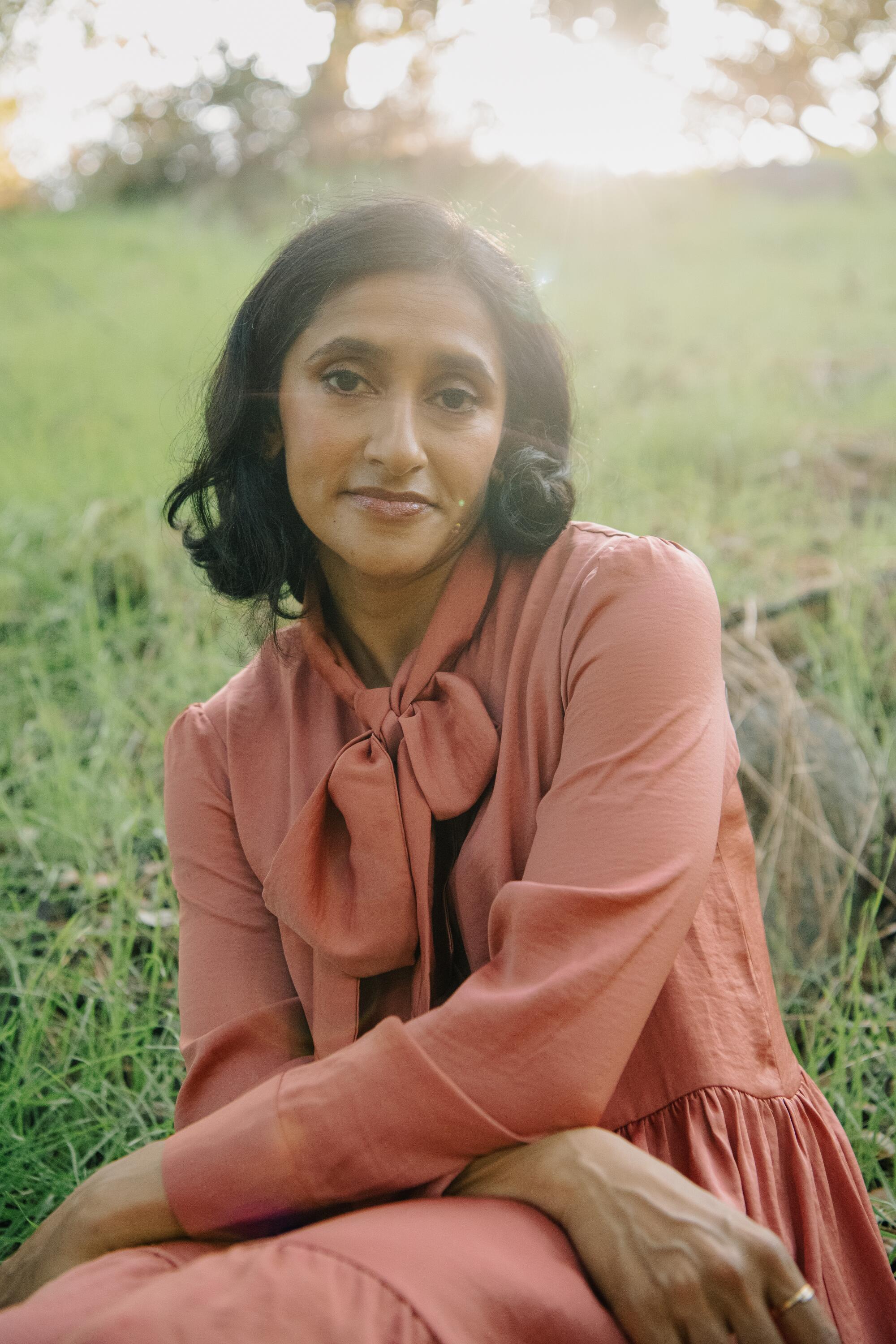 A closeup of a woman in a pinkish dress, sitting in a field.