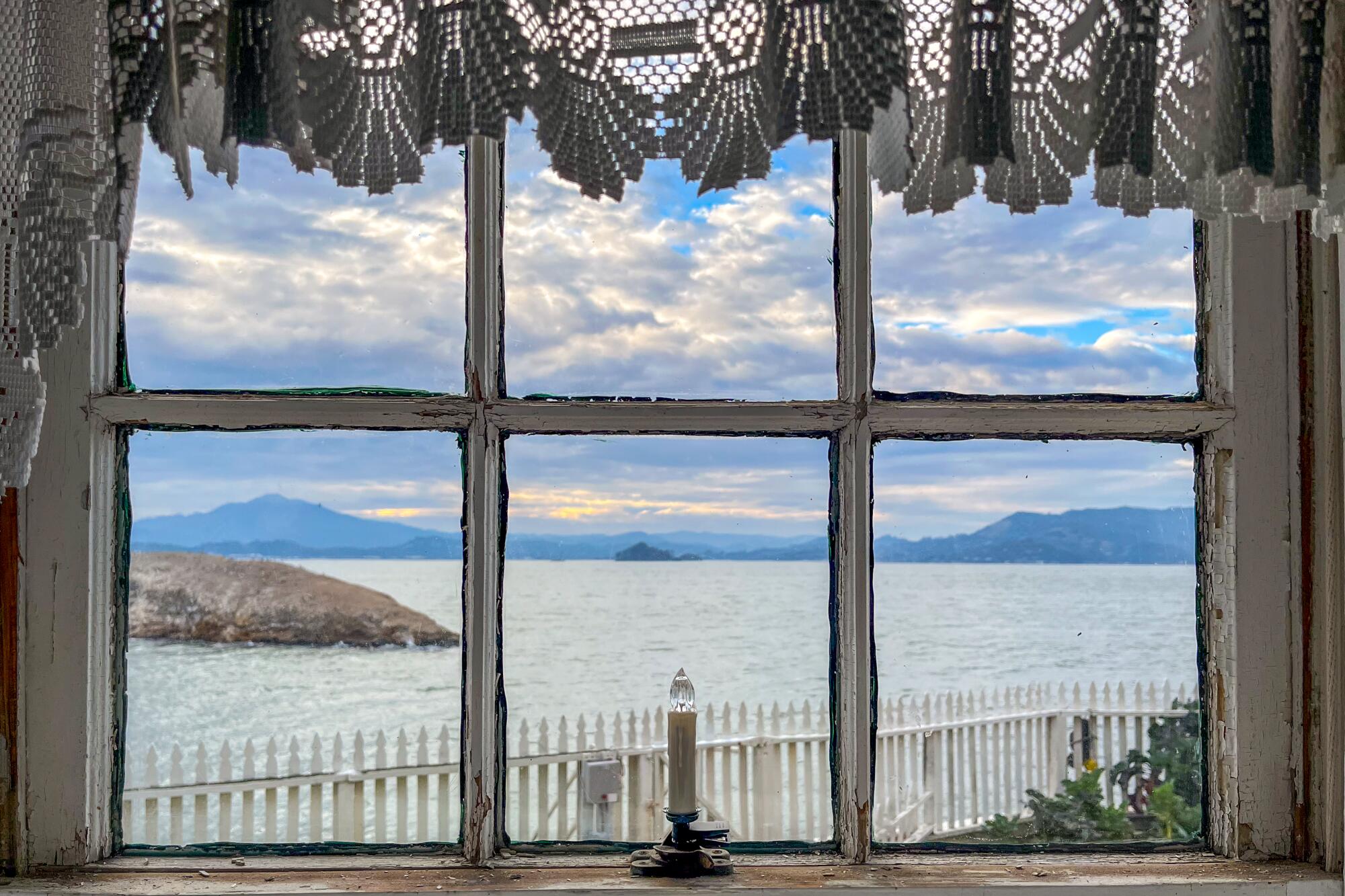 A view through a lace-curtained window of a white picket fence, greenery and the bay.