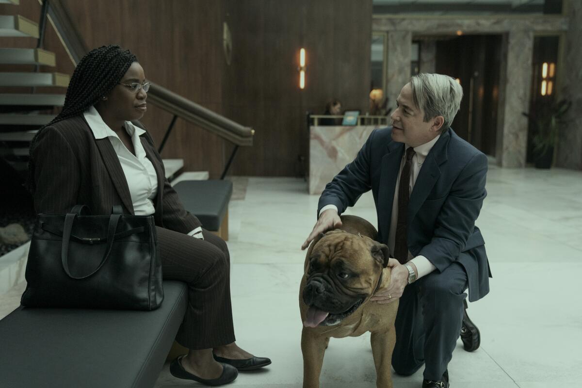 A woman in a suit sits on a bench and talk to a man in a suit who is petting a large brown dog.