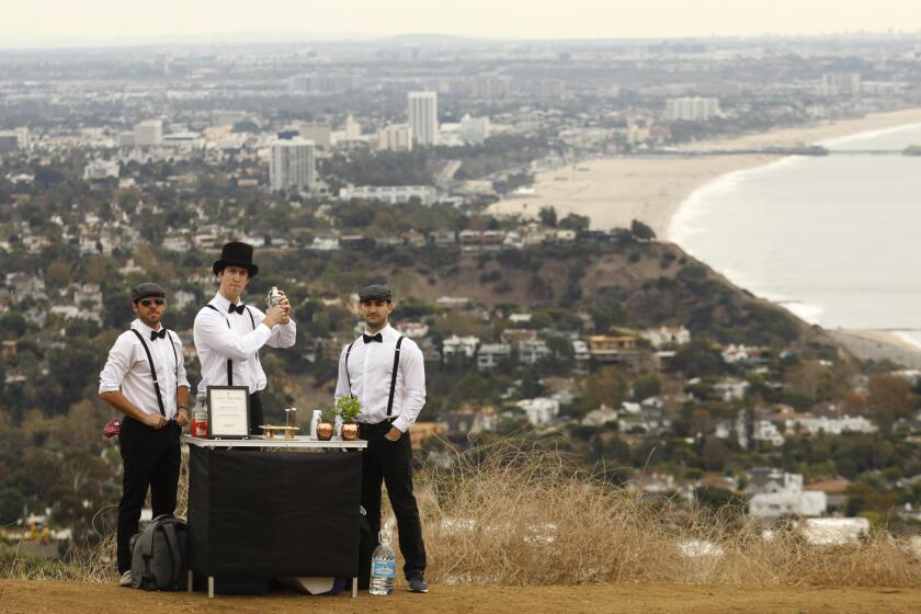 PACIFIC PALISADES, CA - NOVEMBER 30, 2019 - Jack Petros, 28, from left, David Weber, 27, and Dylan Skolnik, 24, members of the Summit Sippers, stand ready to make free drinks for hikers along the Liones Trail in Pacific Palisades on November 30, 2019. Santa Monica Bay and the Santa Monica Pier is viewed in the background. Summit Sippers set up impromptu little bars at random view spots on L.A. hiking trails. They wear bow ties, suspenders and one wears a top hat. They are all avid hikers who enjoy the experience of hiking and than setting up their impromptu watering hole. The drinks are free and they won't even take tips. They are careful not to leave trash behind or block spots where hikers might like to take photos. They try and find picturesque sites along LA’s hiking trails. (Genaro Molina / Los Angeles Times)