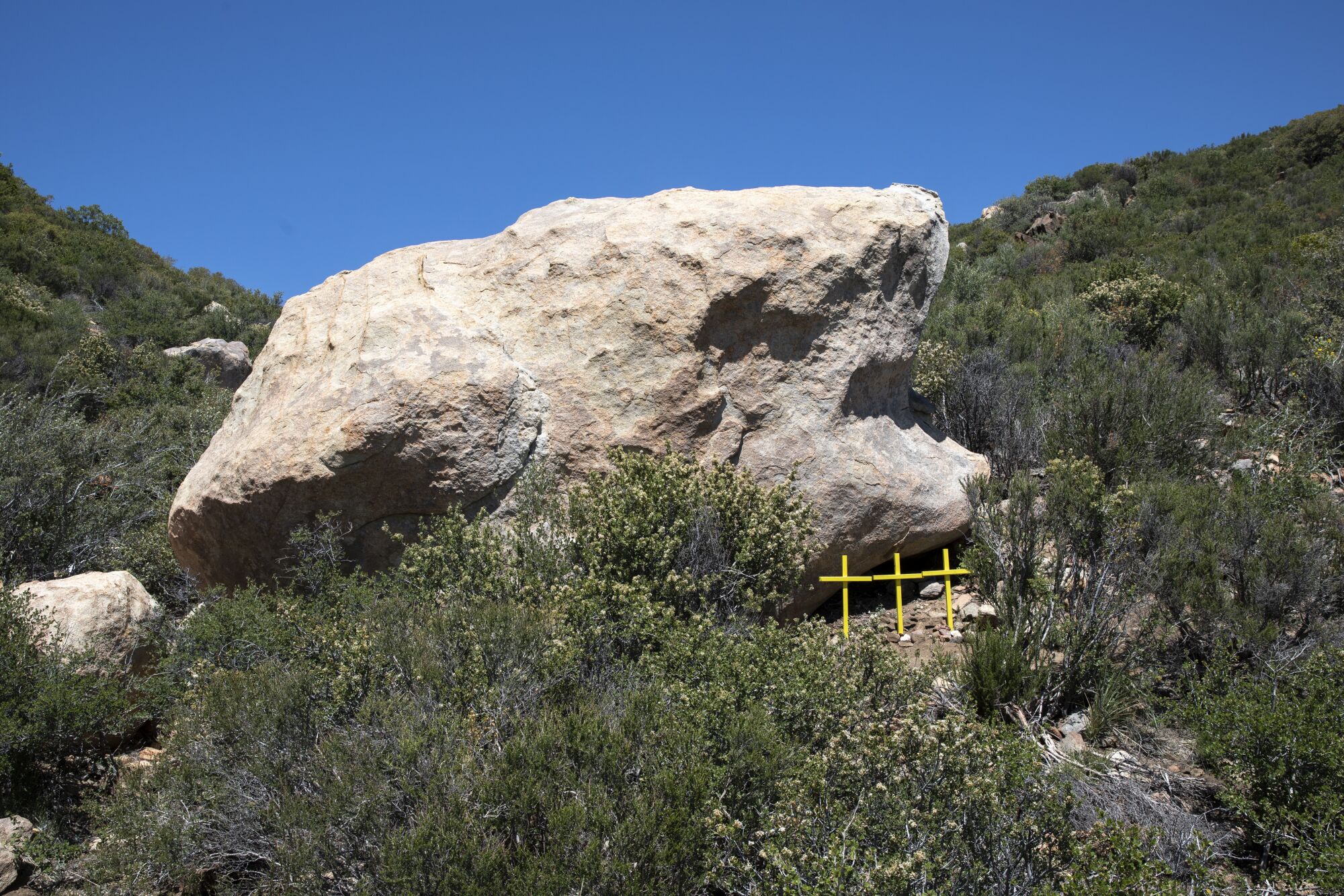 Three yellow crosses planted near the base of a large rock surrounded by shrubs.
