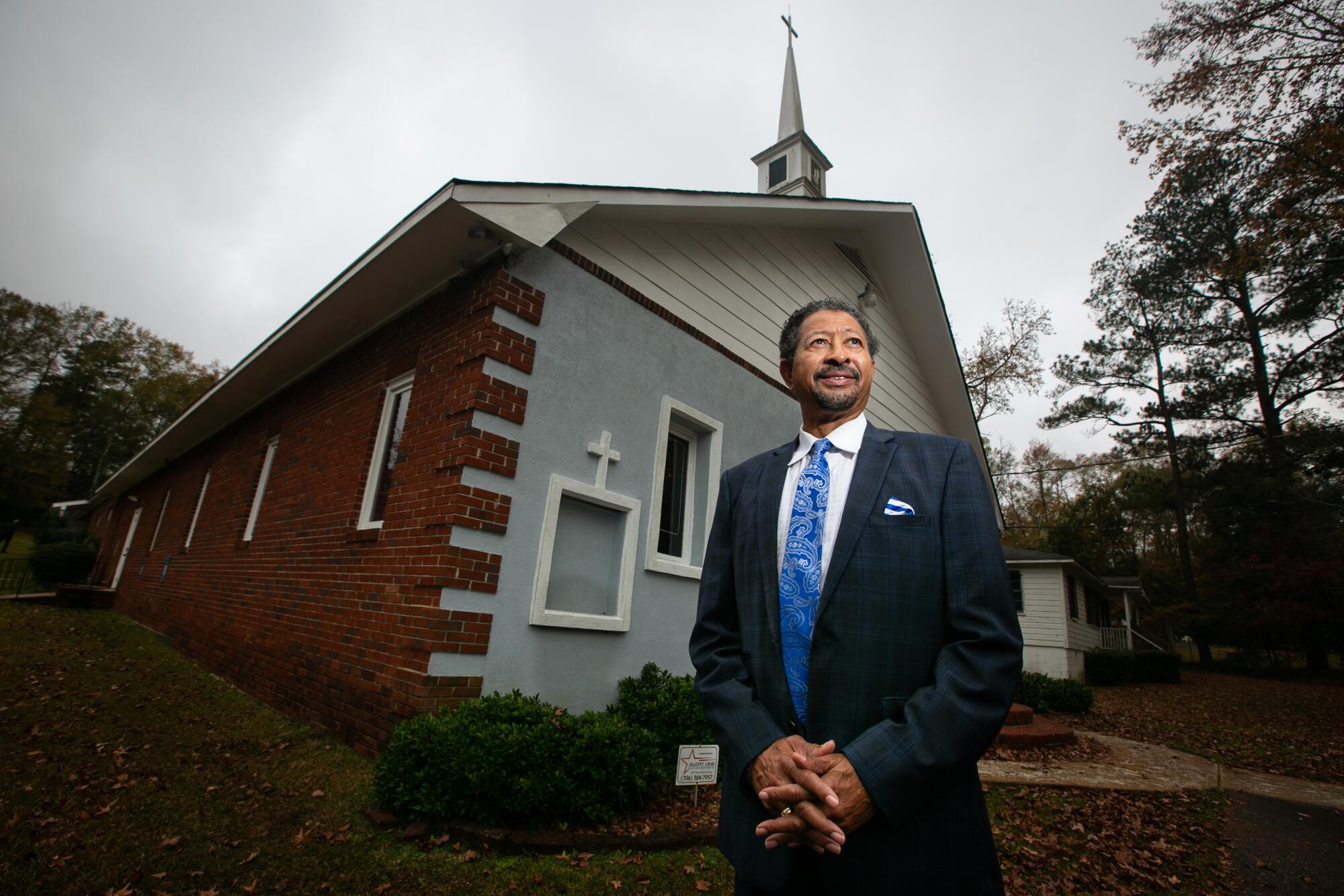  A man in a suit stands with hands clasped in front of a small church building.