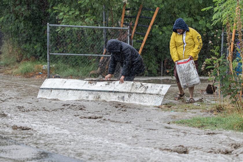 THERMAL, CA - AUGUST 20, 2023: Rafael Nunez, 29, right, with Isabel Ramirez, 23, carries bags of fertilizer to prop up a piece of roofing to try and divert flood waters on 70th Avenue from coming into his mobile home as tropical storm Hilary dumps torrential rain on the area on August 20, 2023 in Thermal, California. (Gina Ferazzi / Los Angeles Times)
