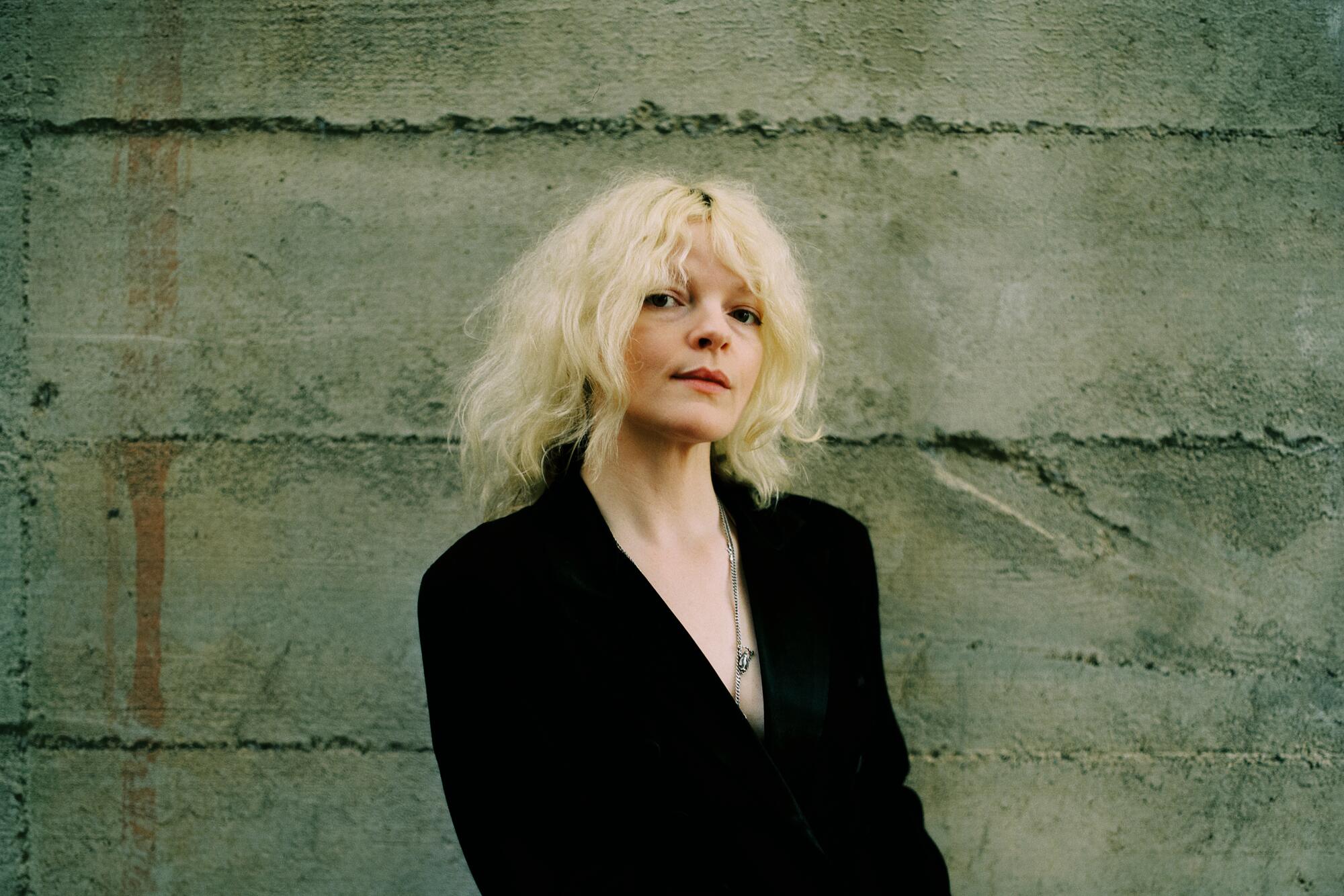 Blond woman in black jacket standing in front of a gray wall