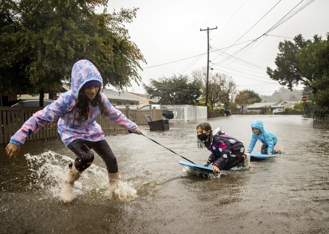 Two children play on boards in floodwaters while one child pulls them
