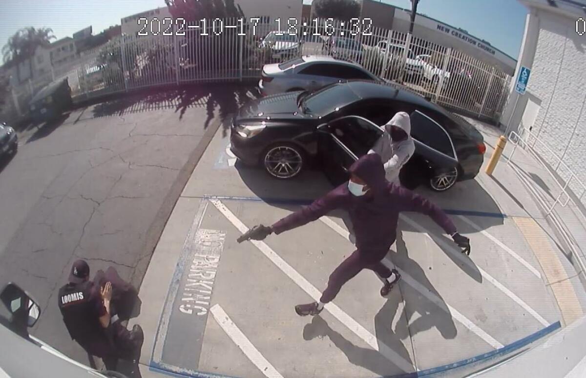 Two men pointing handguns at a guard crouching next to an armored car in a parking lot