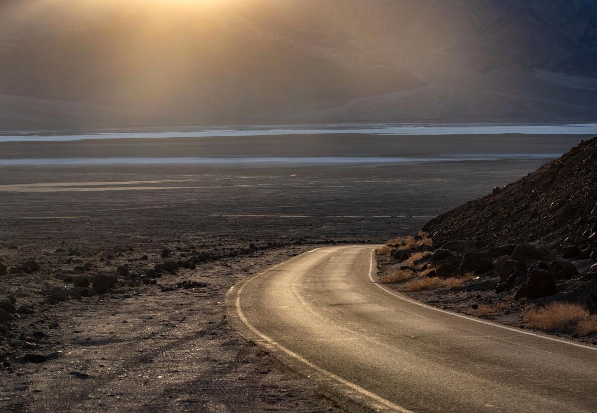 A road curves around a rocky outcrop heading into the setting sun toward a desert basin.