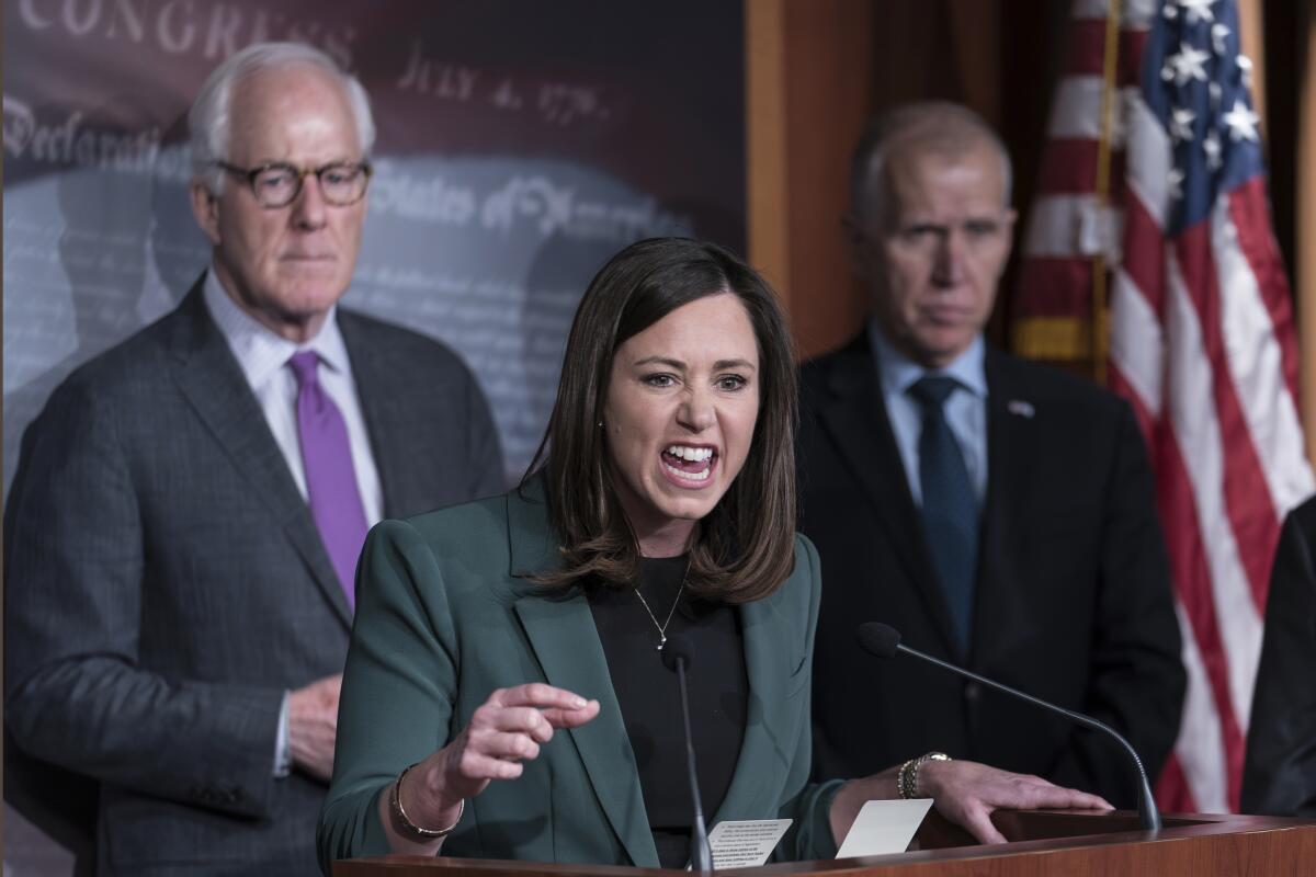 A woman speaking and gesturing, flanked by two men in a Senate briefing room 
