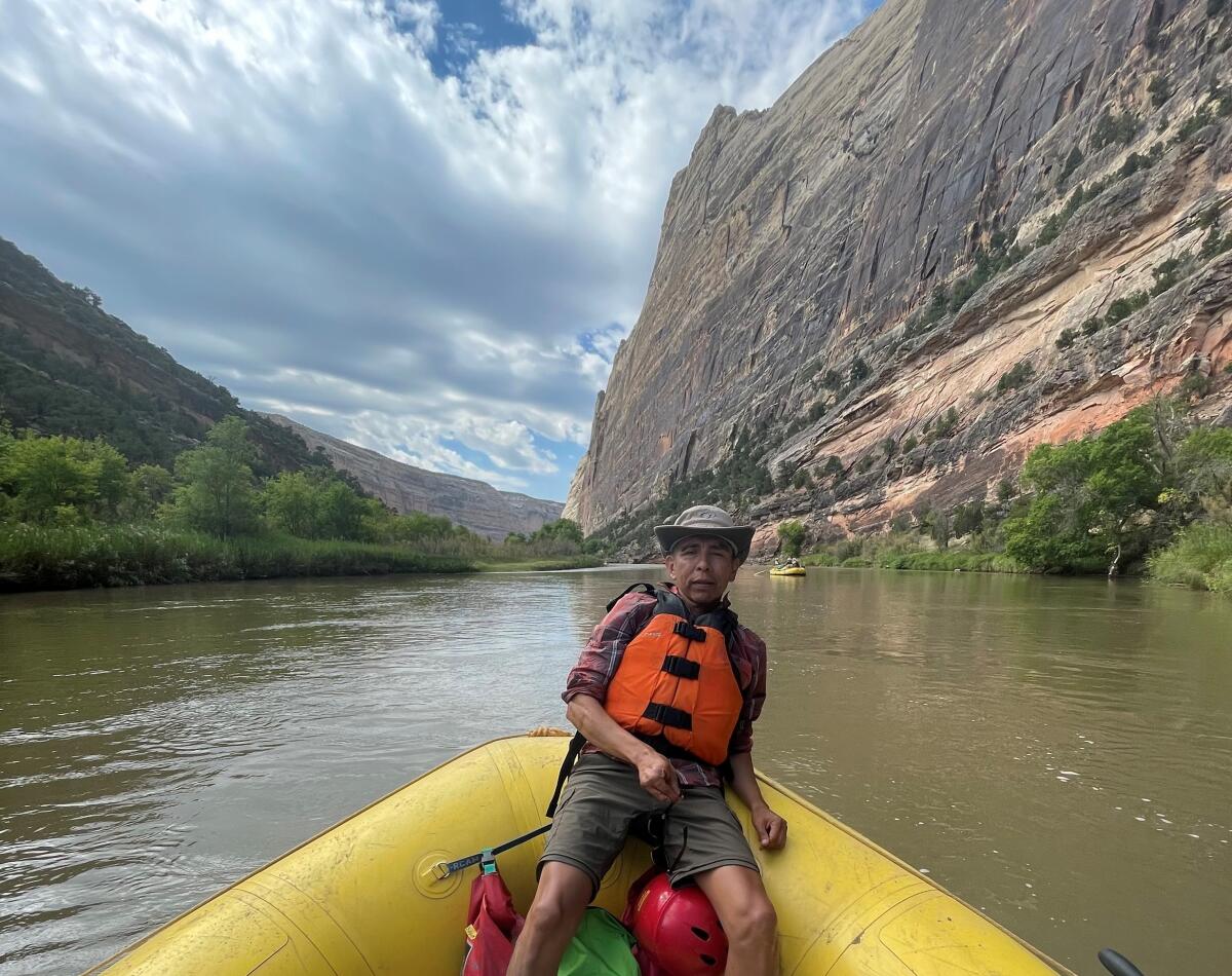 A man in an orange vest and hat leans back in an inflatable boat on a wide river bordered by cliffs and trees.