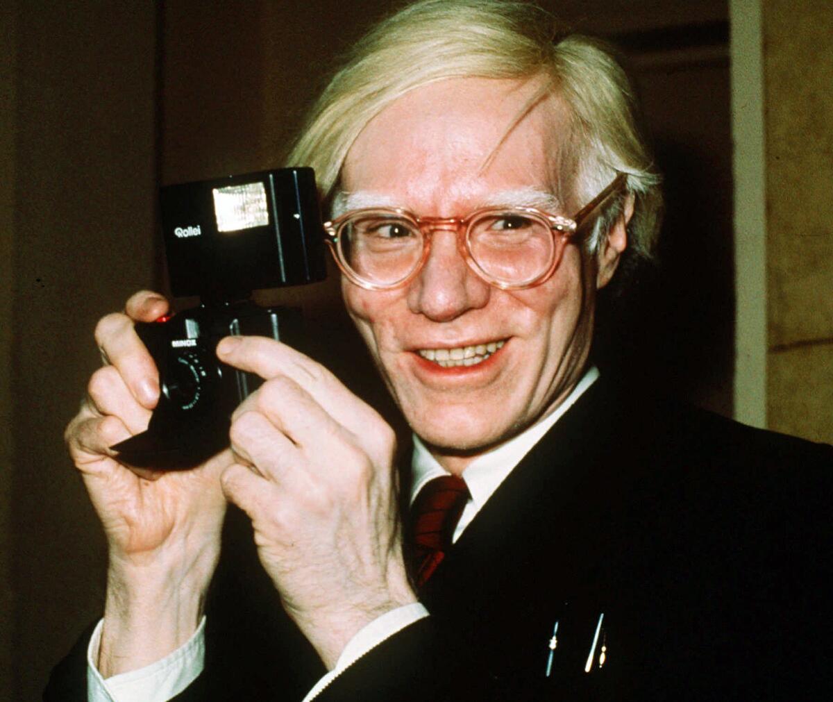 Andy Warhol holds a camera and smiles in 1976