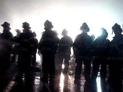 Silhouetted firefighters watch rescue workers look for survivors