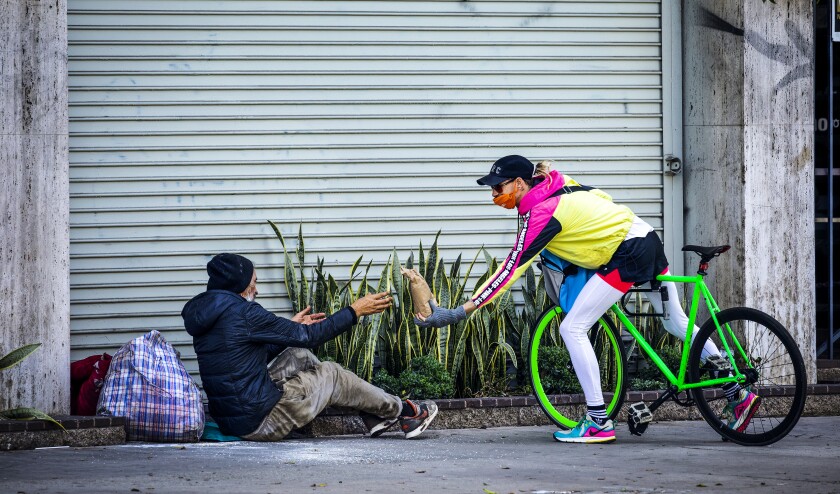 A person on a bicycle reaches out to hand a brown paper bag meal to a man sitting on the sidewalk