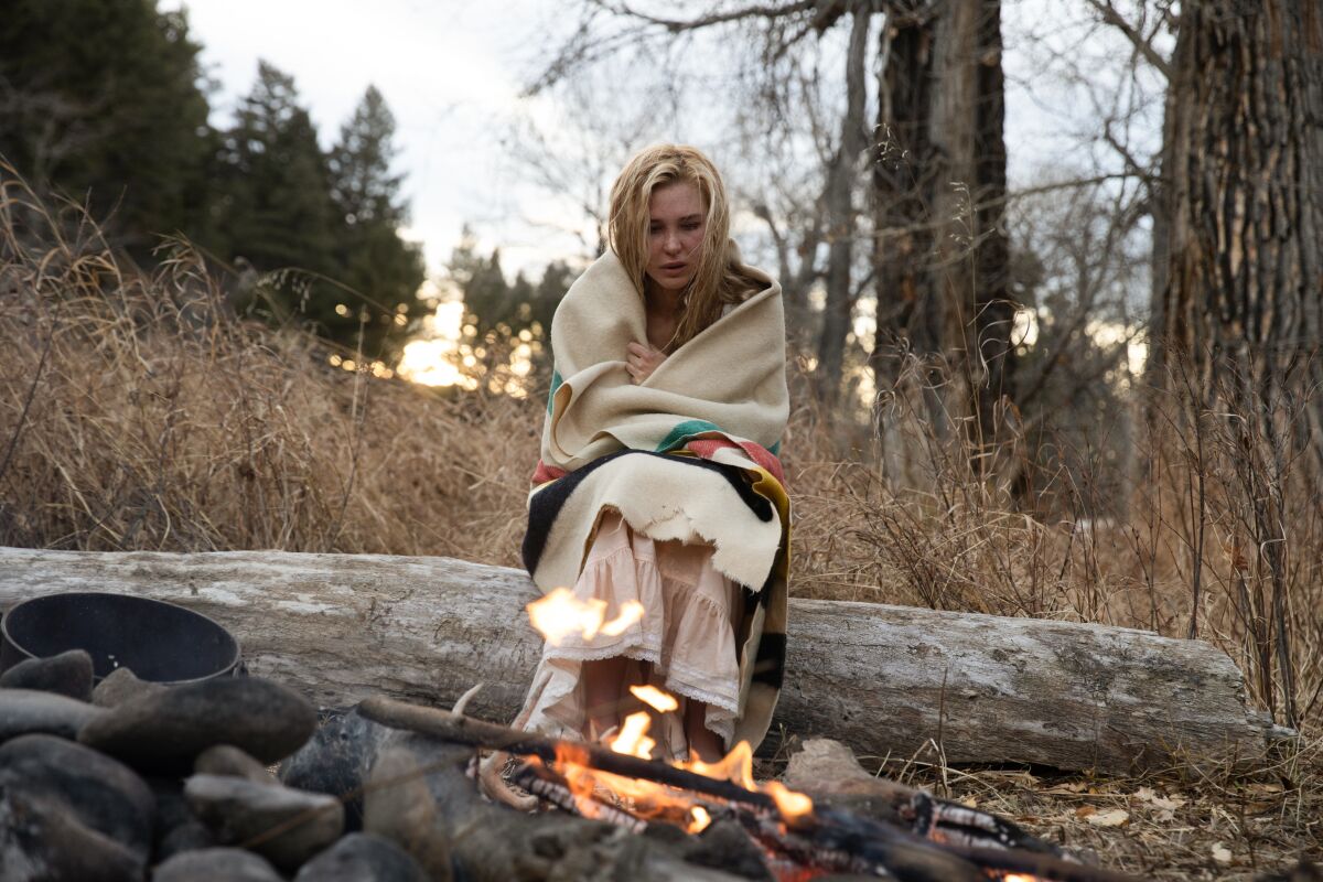 A woman wrapped in a blanket sits on a log by a fire